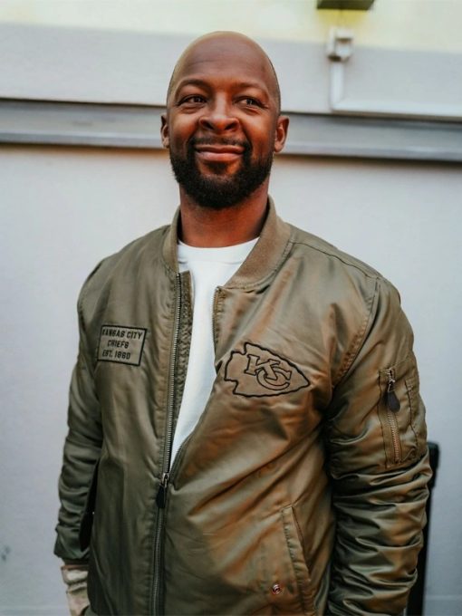 Man wearing a green bomber jacket with a logo, standing against a light-colored wall.